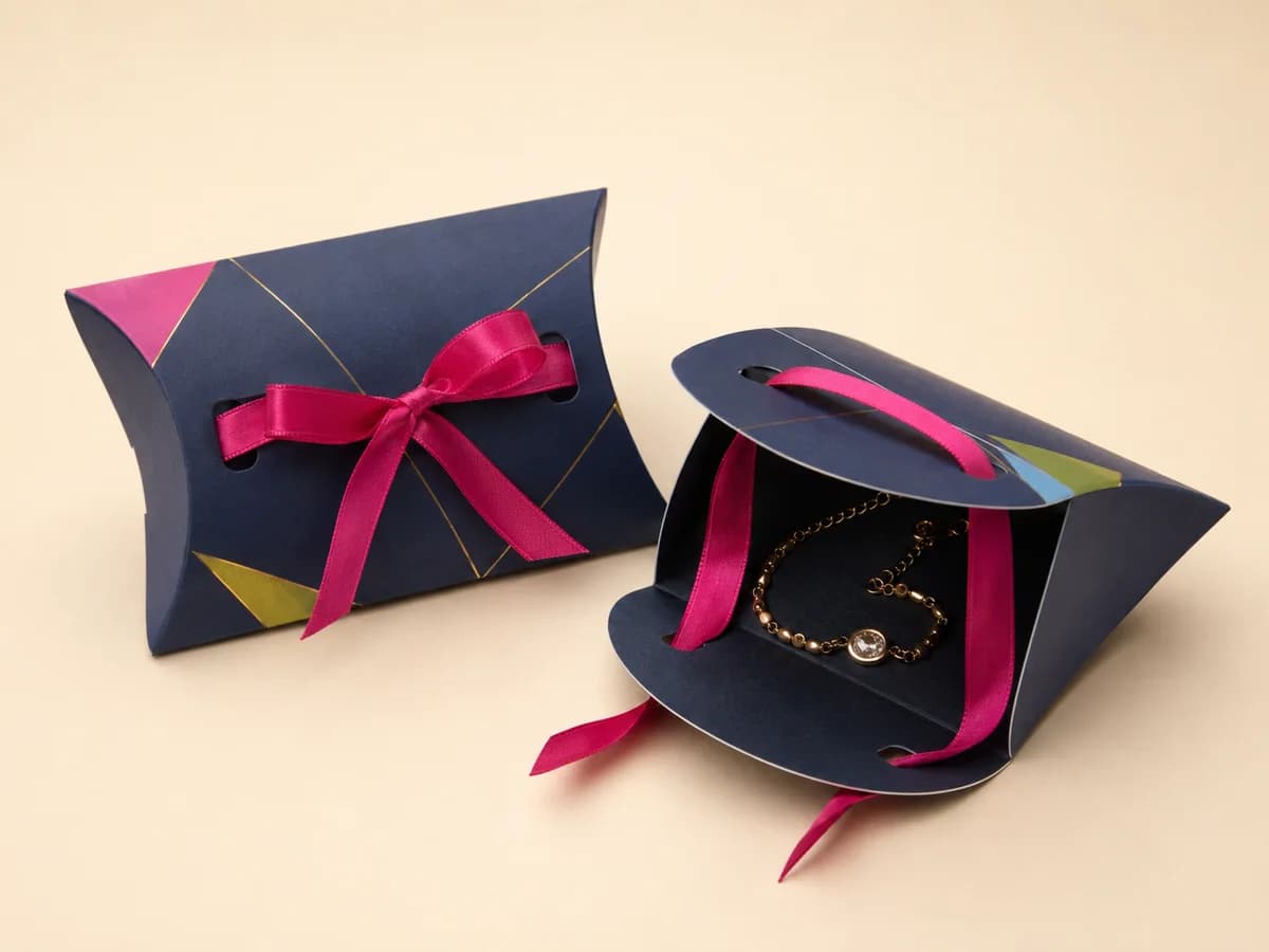 Navy pillow-shaped jewellery box with pink satin ribbon bow, shown closed and open with a bracelet